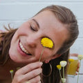 Woman with a yellow flower on her eye, surrounded by flowers and a drink.