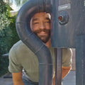 Person peeking out from behind a large metal pipe outdoors