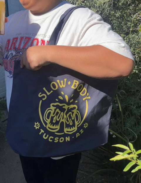 Person holding a blue tote bag with 'Slow Body Tucson, AZ' printed on it.