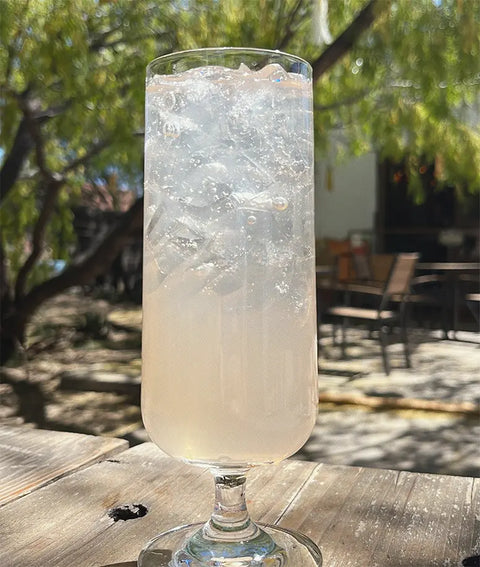 Highball glass with a clear drink and ice cubes on a wooden table outdoors.